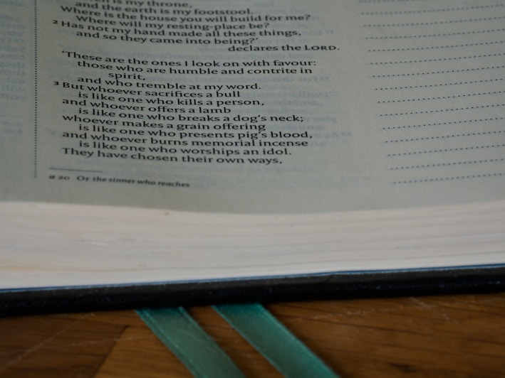 An open book displaying text with a focus on certain passages that discuss spiritual and moral themes. The page is light-colored with black printed text and a teal ribbon bookmark partially visible at the bottom. The book rests on a wooden surface.