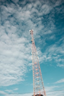 A tall communication tower stands prominently against a partly cloudy sky. The tower has a red and white structure and is equipped with multiple antennas.