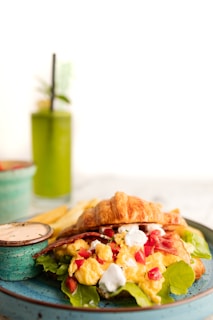 A breakfast croissant sandwich filled with scrambled eggs, red bell pepper pieces, lettuce, and topped with a white creamy sauce. It is accompanied by a side of dipping sauce and fries, served on a turquoise plate. In the background, there is a green smoothie with a straw, garnished with a mint leaf.