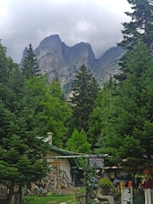 A picturesque mountain scene with towering rocky peaks shrouded in clouds in the background. In the foreground, lush green coniferous trees frame a small, rustic building with a stone foundation and wooden elements. Signage in a foreign script is visible, and various ornamental plants and decorations add charm to the dwelling. The overall setting suggests a peaceful, nature-centered environment.