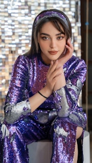 A person with stylish makeup and a serene expression is wearing a shimmering purple sequined outfit and matching headband. The background consists of a blurred array of square tiles, creating an artistic and modern atmosphere.