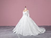 A white wedding dress on a mannequin with intricate lace and floral details, featuring long sleeves and a flowing train. The background is a solid pink wall and the floor is gray with a subtle geometric pattern.