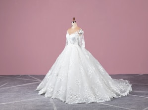 A white wedding dress on a mannequin with intricate lace and floral details, featuring long sleeves and a flowing train. The background is a solid pink wall and the floor is gray with a subtle geometric pattern.