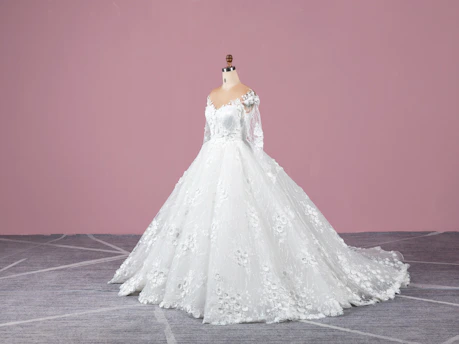 A beautiful white bridal gown displayed on a mannequin with soft pink and beige background.