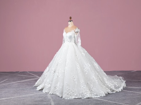 A white wedding dress on a mannequin with intricate lace and floral details, featuring long sleeves and a flowing train. The background is a solid pink wall and the floor is gray with a subtle geometric pattern.