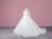 A white wedding dress on a mannequin with intricate lace and floral details, featuring long sleeves and a flowing train. The background is a solid pink wall and the floor is gray with a subtle geometric pattern.