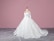 A white wedding dress on a mannequin with intricate lace and floral details, featuring long sleeves and a flowing train. The background is a solid pink wall and the floor is gray with a subtle geometric pattern.