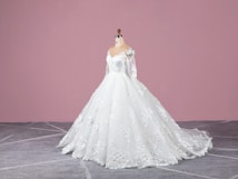 A white wedding dress on a mannequin with intricate lace and floral details, featuring long sleeves and a flowing train. The background is a solid pink wall and the floor is gray with a subtle geometric pattern.