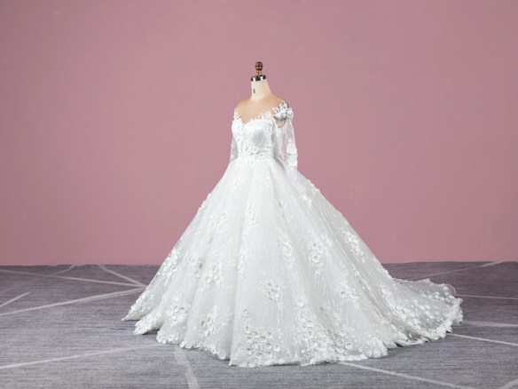 A white wedding dress on a mannequin with intricate lace and floral details, featuring long sleeves and a flowing train. The background is a solid pink wall and the floor is gray with a subtle geometric pattern.