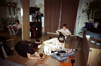 A cozy indoor setting with three people engaged in a musical activity. One person is playing an electric guitar while another observes closely. A third person is seated in the background near a curtain. The room has wooden flooring, and there are various objects like a bookshelf, plants, and a mirrored wardrobe visible.