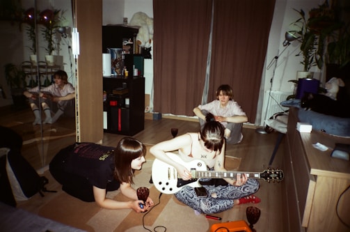 A cozy indoor setting with three people engaged in a musical activity. One person is playing an electric guitar while another observes closely. A third person is seated in the background near a curtain. The room has wooden flooring, and there are various objects like a bookshelf, plants, and a mirrored wardrobe visible.
