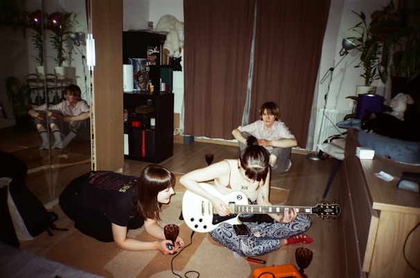A cozy indoor setting with three people engaged in a musical activity. One person is playing an electric guitar while another observes closely. A third person is seated in the background near a curtain. The room has wooden flooring, and there are various objects like a bookshelf, plants, and a mirrored wardrobe visible.