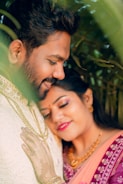 A joyful and romantic couple is embracing amidst lush greenery. The woman is resting her head gently on the man's chest with a serene smile, wearing a colorful, embroidered saree and adorned with jewelry. The man is smiling softly and clad in a light traditional outfit, their warmth and affection palpable. The foliage around them adds a vibrant, natural frame to the intimate moment.
