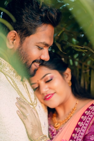 A joyful and romantic couple is embracing amidst lush greenery. The woman is resting her head gently on the man's chest with a serene smile, wearing a colorful, embroidered saree and adorned with jewelry. The man is smiling softly and clad in a light traditional outfit, their warmth and affection palpable. The foliage around them adds a vibrant, natural frame to the intimate moment.