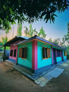 A brightly colored house with turquoise, pink, and yellow painted walls, featuring decorative windows with wooden shutters. The house is surrounded by lush palm trees and other vegetation under a clear sky. There are some mats or clothes laying on the ground near the house, and large tree branches with leaves frame the upper part of the image.