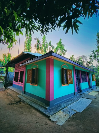 A brightly colored house with turquoise, pink, and yellow painted walls, featuring decorative windows with wooden shutters. The house is surrounded by lush palm trees and other vegetation under a clear sky. There are some mats or clothes laying on the ground near the house, and large tree branches with leaves frame the upper part of the image.