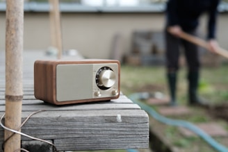 A vintage-style radio with a wooden finish is placed on a wooden surface in an outdoor setting. A person in the background, blurred, appears to be gardening or tending to plants.