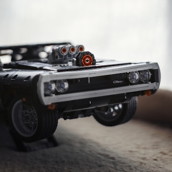A detailed close-up view of a LEGO model car, resembling a classic muscle car, with a powerful engine protruding from the hood. The model displays intricate details like headlights and a shiny, polished grille. The background is softly blurred, emphasizing the model's craftsmanship.