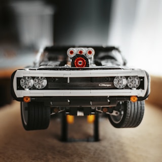 A detailed and realistic LEGO model of a classic car, complete with intricate features such as a front grille, headlights, and prominent front tires. The model is positioned on a stand, showcasing its structure and build.