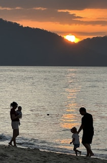 A serene beach sunset with a family in silhouette. The sun is setting behind distant hills, casting an orange glow over the calm ocean. A woman holds a baby, while a man walks hand in hand with a child along the sandy shore.