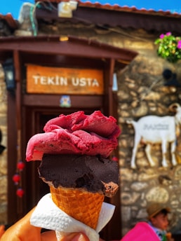 A hand is holding a cone with two scoops of ice cream, one dark brown and one dark pink in color. In the background, there is a sign reading 'Tekin Usta' mounted on a rustic building with stone walls. There's also a white decorative piece resembling an animal, possibly a goat, along with some flowers and a person wearing a hat on the right side.