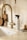 A minimalistic kitchen scene with a modern brass-colored faucet and a countertop. A spray bottle labeled 'all-purpose cleaner' is placed next to a wooden brush with a handle. A small, transparent vase with dried flowers and a set of wooden kitchen boards add a touch of decor. The colors are warm and neutral, creating a cozy and clean atmosphere.
