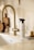A minimalistic kitchen scene with a modern brass-colored faucet and a countertop. A spray bottle labeled 'all-purpose cleaner' is placed next to a wooden brush with a handle. A small, transparent vase with dried flowers and a set of wooden kitchen boards add a touch of decor. The colors are warm and neutral, creating a cozy and clean atmosphere.