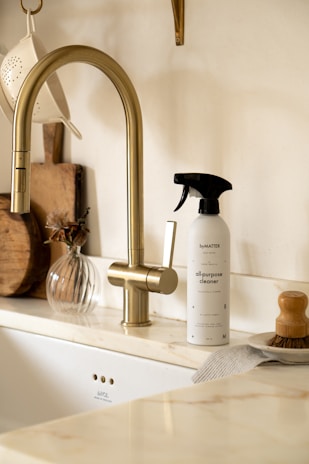 A minimalistic kitchen scene with a modern brass-colored faucet and a countertop. A spray bottle labeled 'all-purpose cleaner' is placed next to a wooden brush with a handle. A small, transparent vase with dried flowers and a set of wooden kitchen boards add a touch of decor. The colors are warm and neutral, creating a cozy and clean atmosphere.
