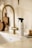 A minimalistic kitchen scene with a modern brass-colored faucet and a countertop. A spray bottle labeled 'all-purpose cleaner' is placed next to a wooden brush with a handle. A small, transparent vase with dried flowers and a set of wooden kitchen boards add a touch of decor. The colors are warm and neutral, creating a cozy and clean atmosphere.