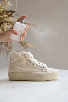 A hand holding a spray bottle labeled 'another day' is spraying mist over a beige high-top sneaker. The sneaker is positioned on a smooth, light-colored surface, with dried plant stems in the background.
