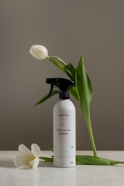 A bottle of all-purpose cleaner labeled 'byMATTER' is set on a light surface. Surrounding the bottle are two white tulips, one leaned against the bottle and another resting on the surface. The backdrop is an earthy gray, creating a calm and minimalist aesthetic.