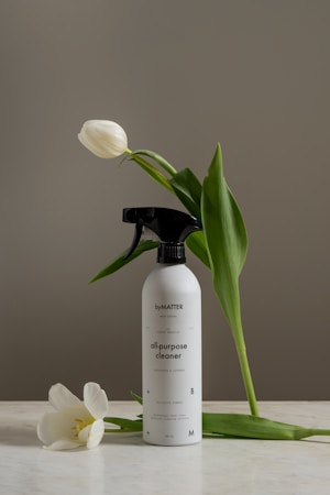 A bottle of all-purpose cleaner labeled 'byMATTER' is set on a light surface. Surrounding the bottle are two white tulips, one leaned against the bottle and another resting on the surface. The backdrop is an earthy gray, creating a calm and minimalist aesthetic.