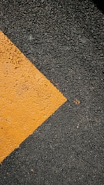 A textured asphalt surface with a prominent diagonal yellow paint stripe running across it. The paint appears slightly worn, contrasting against the dark, rough texture of the asphalt.