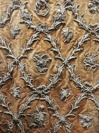 Intricate silver embroidery designs create a repeating pattern on a rich, textured brown fabric. The motifs include floral elements and ornate scrolls, giving it an elegant and luxurious appearance.