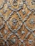 Intricate silver embroidery designs create a repeating pattern on a rich, textured brown fabric. The motifs include floral elements and ornate scrolls, giving it an elegant and luxurious appearance.