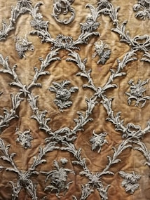 Intricate silver embroidery designs create a repeating pattern on a rich, textured brown fabric. The motifs include floral elements and ornate scrolls, giving it an elegant and luxurious appearance.