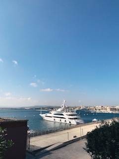 A large, sleek white yacht is docked beside a concrete pier on a calm, blue sea under a clear, sunny sky. In the background, there are more boats and a cityscape with low-rise buildings along the coastline.