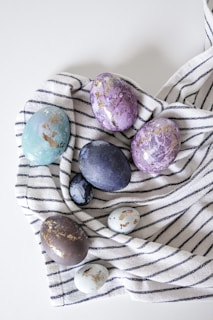 A collection of decorated eggs rests on a striped cloth. The eggs are artistically adorned with colors like purple, blue, and hints of gold leaf detailing. The cloth underneath has a pattern of dark navy stripes against a white background.