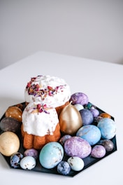A decorative display featuring a baked cake with white icing and flower petals, surrounded by a variety of colored Easter eggs on a black plate. The eggs are painted in shades of blue, purple, gold, and cream, with marbled patterns and metallic finishes.