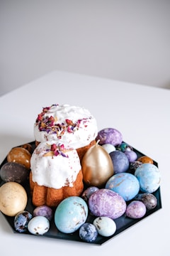 A decorative display featuring a baked cake with white icing and flower petals, surrounded by a variety of colored Easter eggs on a black plate. The eggs are painted in shades of blue, purple, gold, and cream, with marbled patterns and metallic finishes.