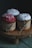 Three cupcakes are decorated with colorful frosting and sprinkles, each wrapped in brown paper and tied with string, placed on a wooden pedestal. The lighting is dim, highlighting the textures and colors of the cupcakes.