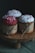 Three cupcakes are decorated with colorful frosting and sprinkles, each wrapped in brown paper and tied with string, placed on a wooden pedestal. The lighting is dim, highlighting the textures and colors of the cupcakes.