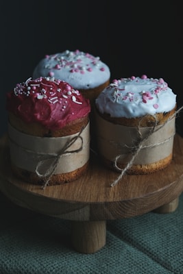 Three cupcakes are decorated with colorful frosting and sprinkles, each wrapped in brown paper and tied with string, placed on a wooden pedestal. The lighting is dim, highlighting the textures and colors of the cupcakes.