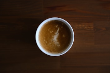 A cup of coffee with a frothy surface sits on a dark wooden table. The coffee's crema forms irregular patterns on the surface, and the overall atmosphere is warm and inviting.