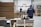 A warehouse setting with workers wearing blue jackets moving stacks of cardboard using pallet jacks. Large piles of cardboard and boxes are organized in the space, suggesting an industrial or shipping environment. The warehouse has a high ceiling and several industrial doors.