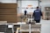 A warehouse setting with workers wearing blue jackets moving stacks of cardboard using pallet jacks. Large piles of cardboard and boxes are organized in the space, suggesting an industrial or shipping environment. The warehouse has a high ceiling and several industrial doors.
