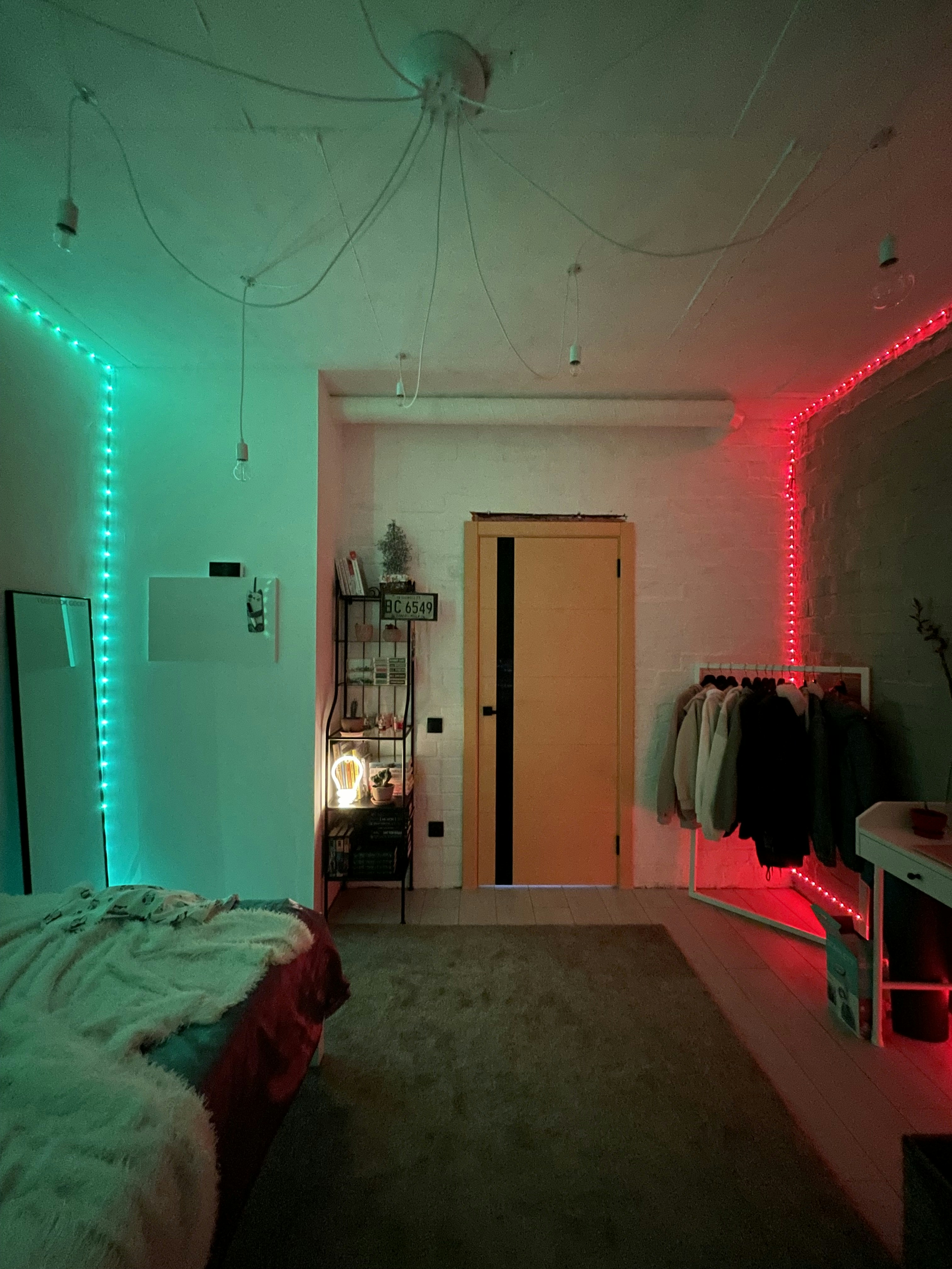 A cozy room with ambient lighting, featuring green and red LED lights along the walls. The room includes a bed with a plush blanket, a shelf with books and decorative items, a clothing rack with jackets, and a door in the center. The atmosphere is warm and inviting, with a minimalist yet stylish decor.