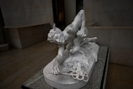 A detailed marble sculpture of a mythological figure, likely a greco-roman god or hero, crouching with wings on the head. The figure appears muscular with fine detailing on the hair and drapery. The sculpture is set on a marble base in an empty, dimly lit gallery.