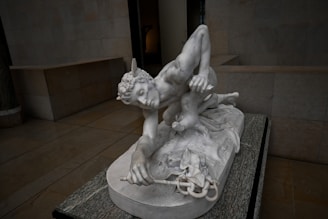 A detailed marble sculpture of a mythological figure, likely a greco-roman god or hero, crouching with wings on the head. The figure appears muscular with fine detailing on the hair and drapery. The sculpture is set on a marble base in an empty, dimly lit gallery.