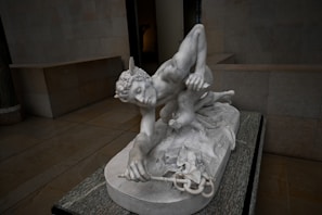 A detailed marble sculpture of a mythological figure, likely a greco-roman god or hero, crouching with wings on the head. The figure appears muscular with fine detailing on the hair and drapery. The sculpture is set on a marble base in an empty, dimly lit gallery.